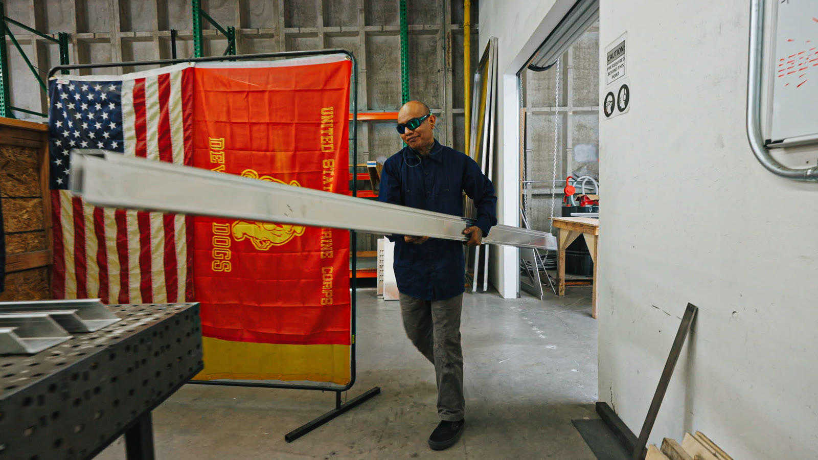 A worker holding a long rectangular silver tube beside a trailer and two hanging flags.