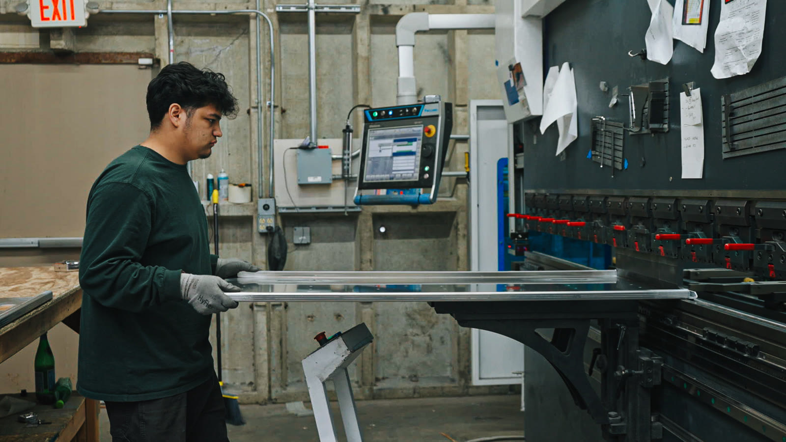 A worker holding a long rectangular silver frame in front of the press-brake control console.