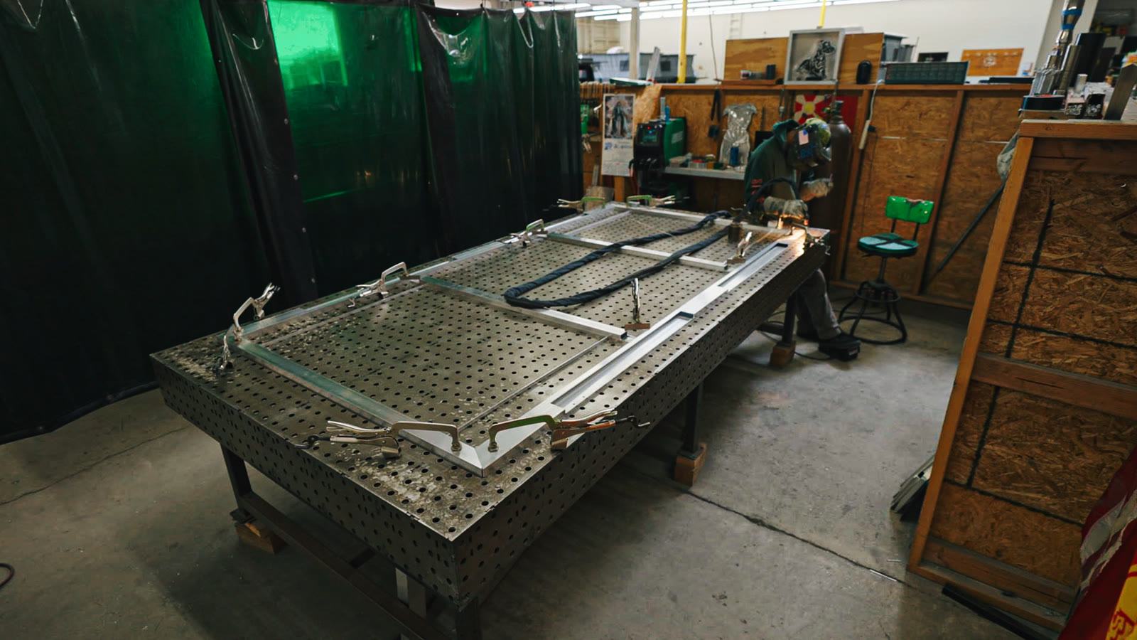 A long silver frame sits diagonally across the perforated table near the welding curtains.