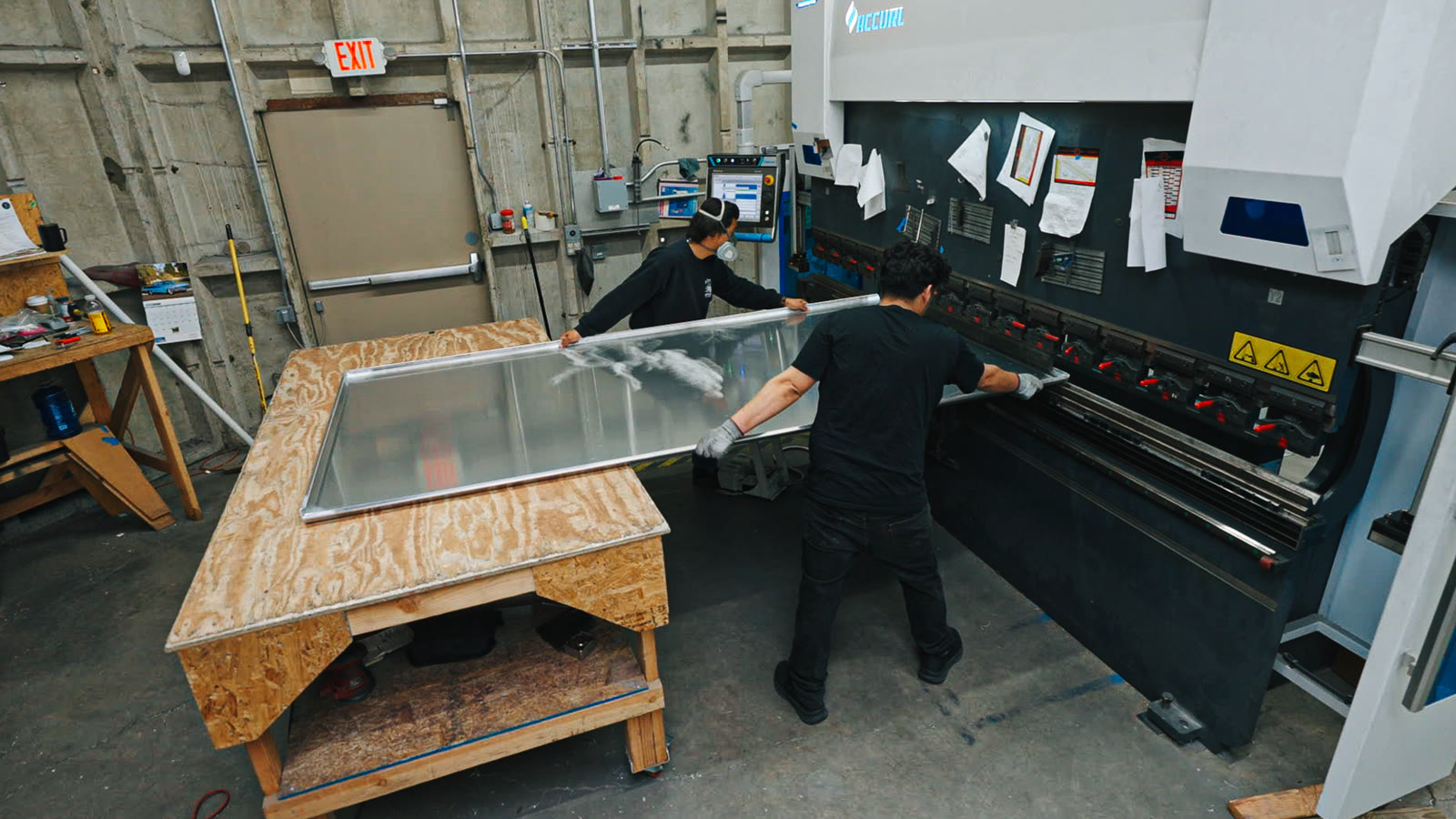 A worker guides a broad silver panel beside the bending machine and plywood workbench.