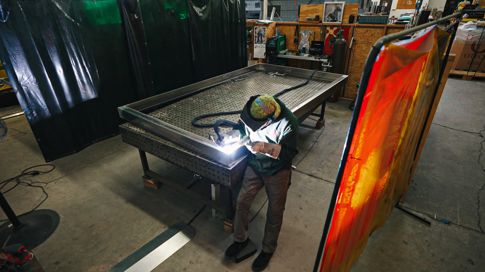 A helmeted technician welding near the corner of a rectangular frame on the perforated worktable.