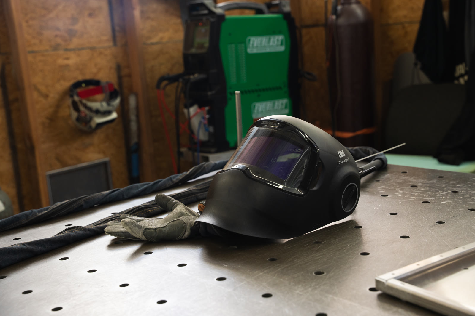 A dark welding helmet and gloves resting on the perforated fabrication table.