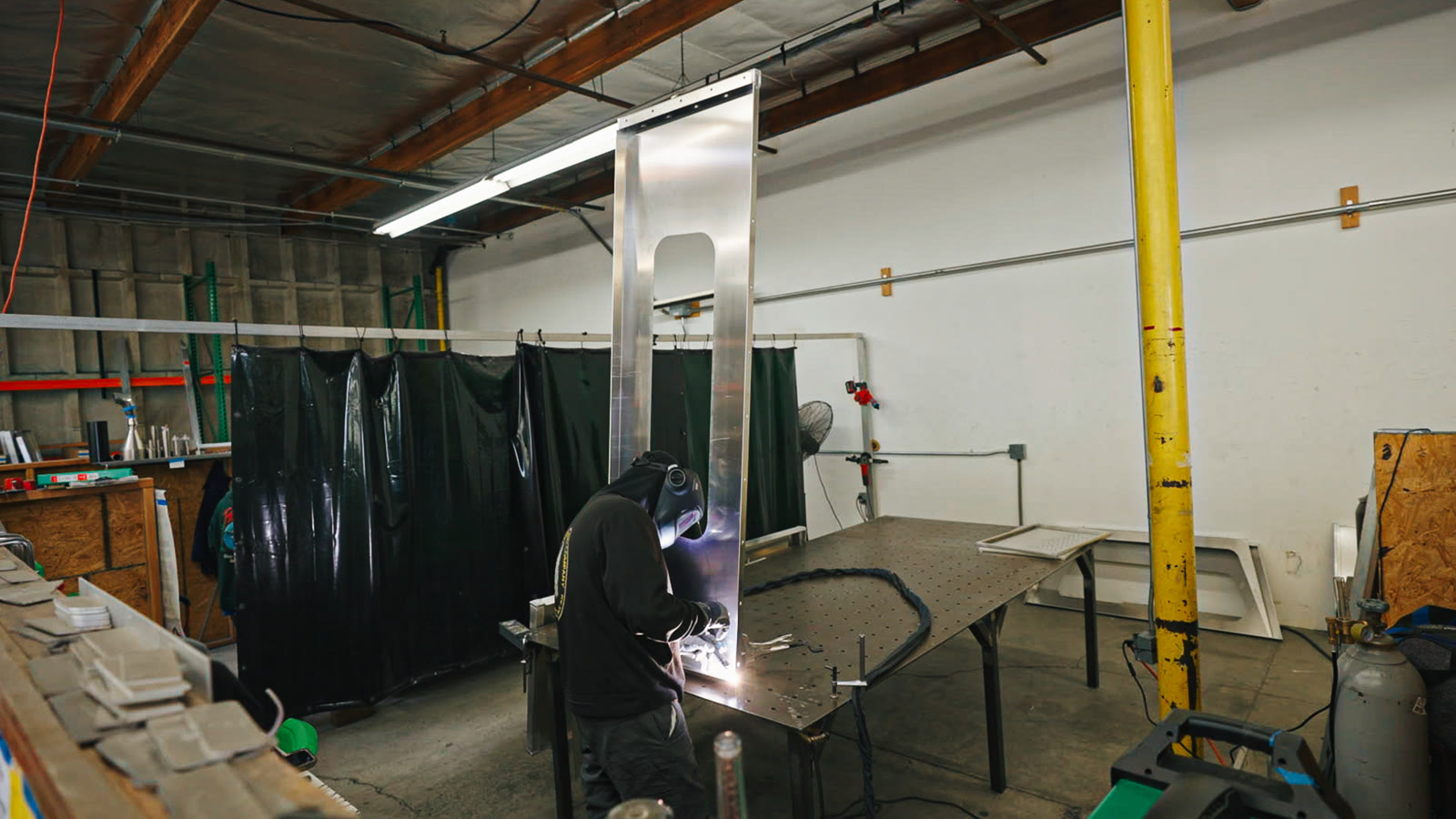 Upright silver frames, green welding curtains, and technicians working across the fabrication area.