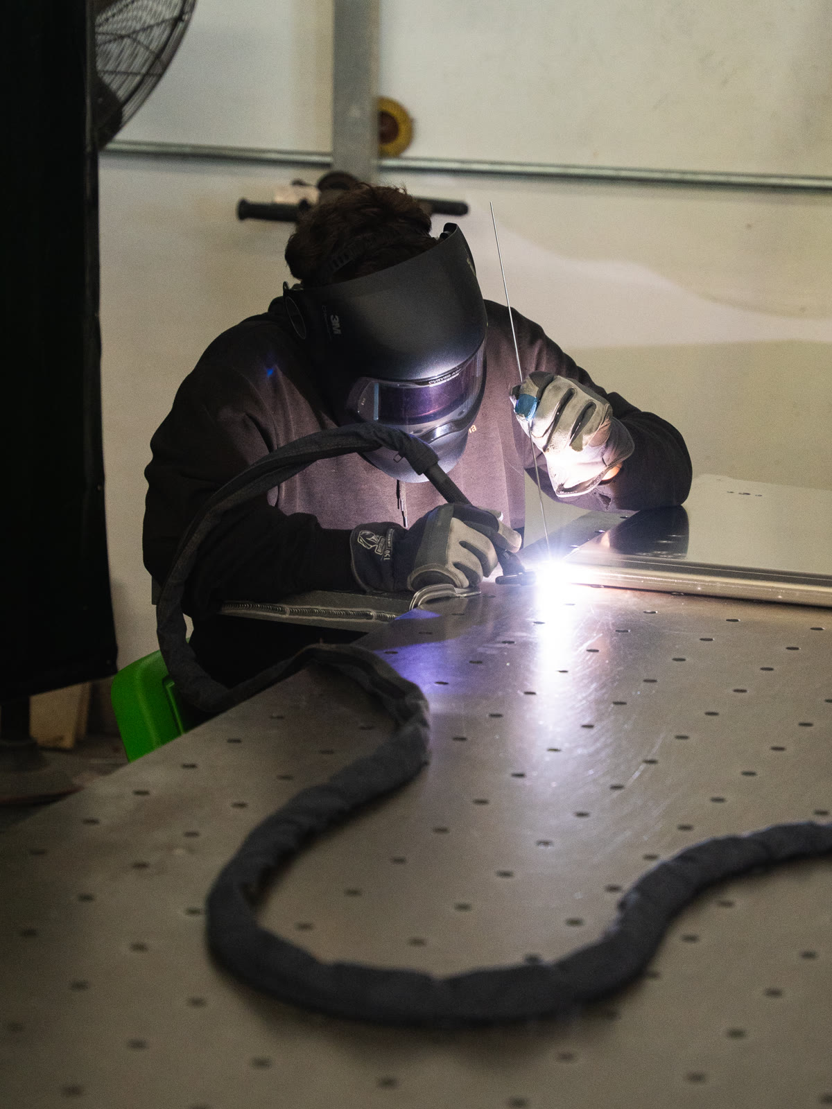 A helmeted technician leans over a metal assembly as a bright welding arc glows.