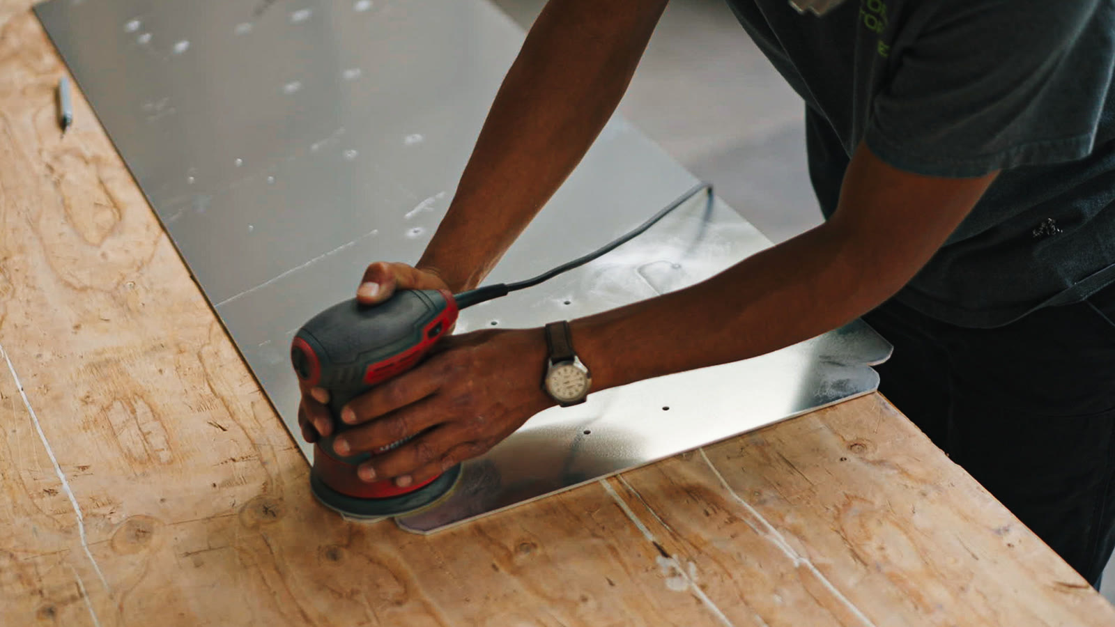 Close view of a worker using a round power sander on a silver panel atop plywood.