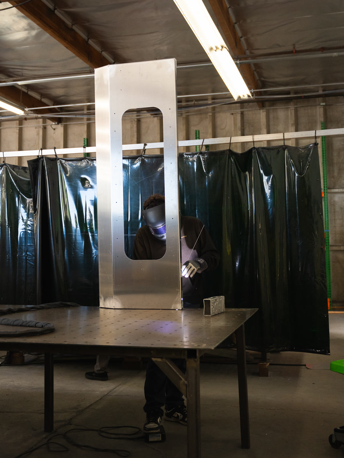 A tall silver rectangular frame stands on a wheeled table in front of dark welding curtains.