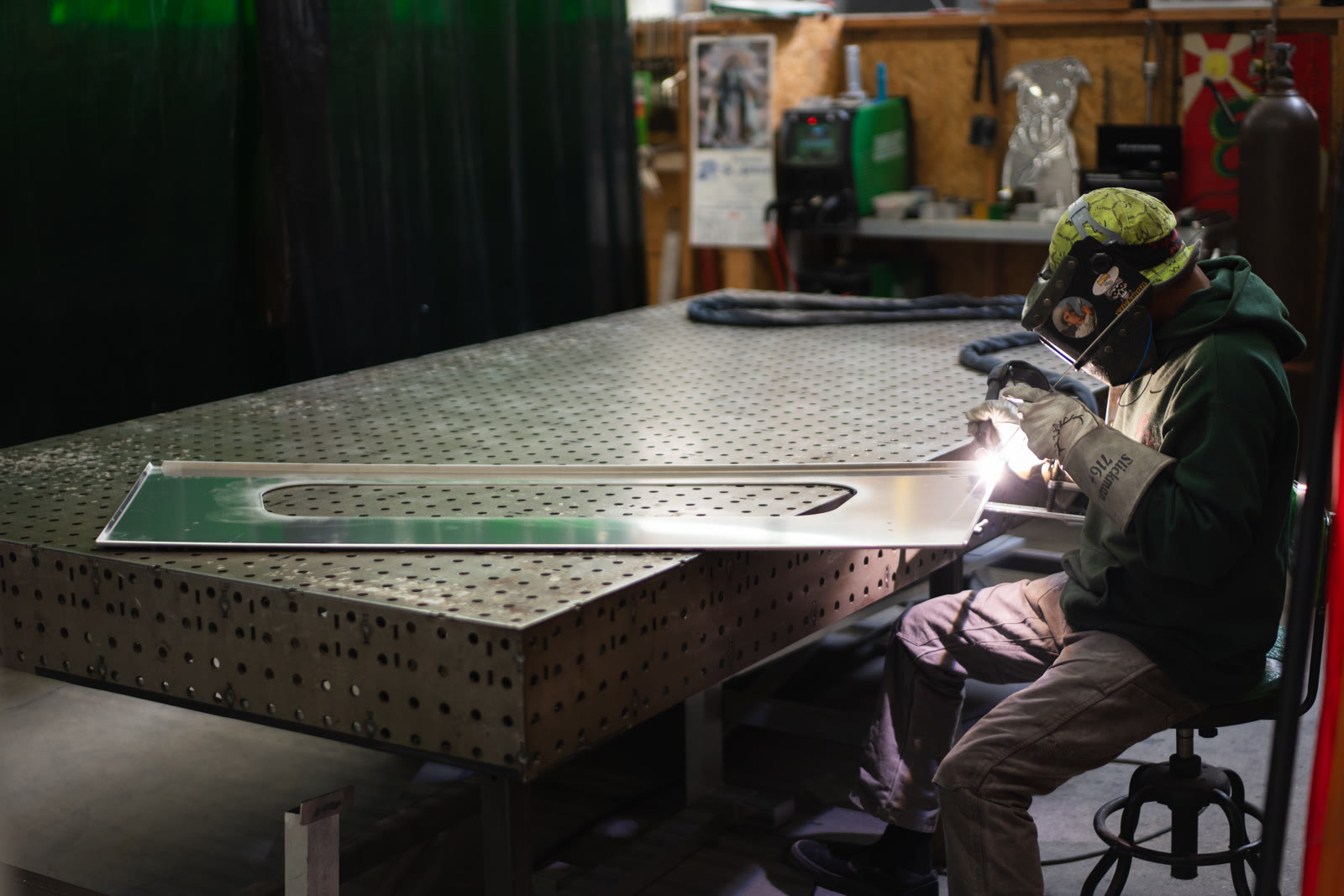 A helmeted technician seated at the corner of a large perforated table while welding a silver frame.
