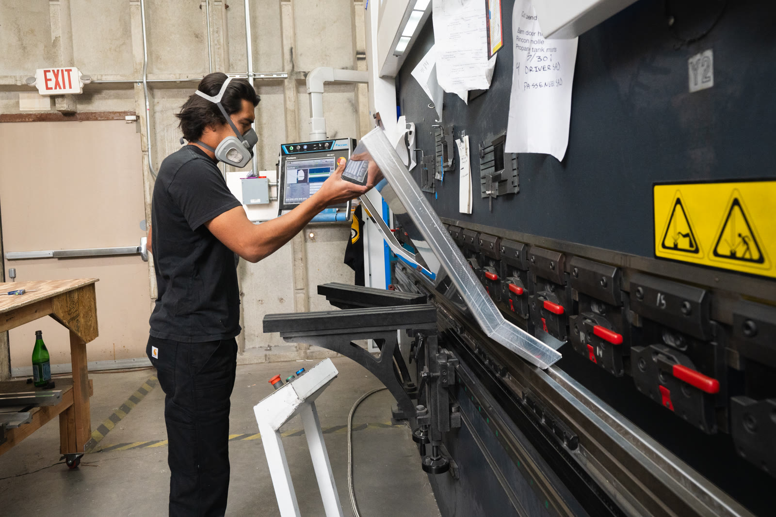 A worker holds a long narrow silver piece beside the controls of the large bending machine.
