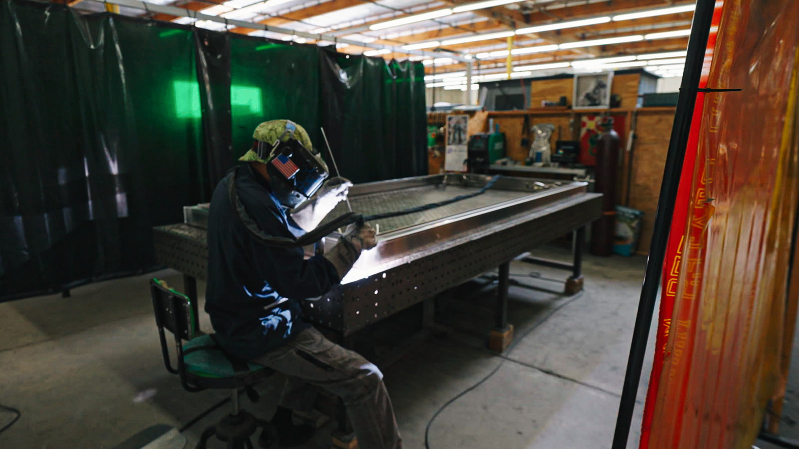 A helmeted technician seated beside the perforated fabrication table while welding a silver frame.
