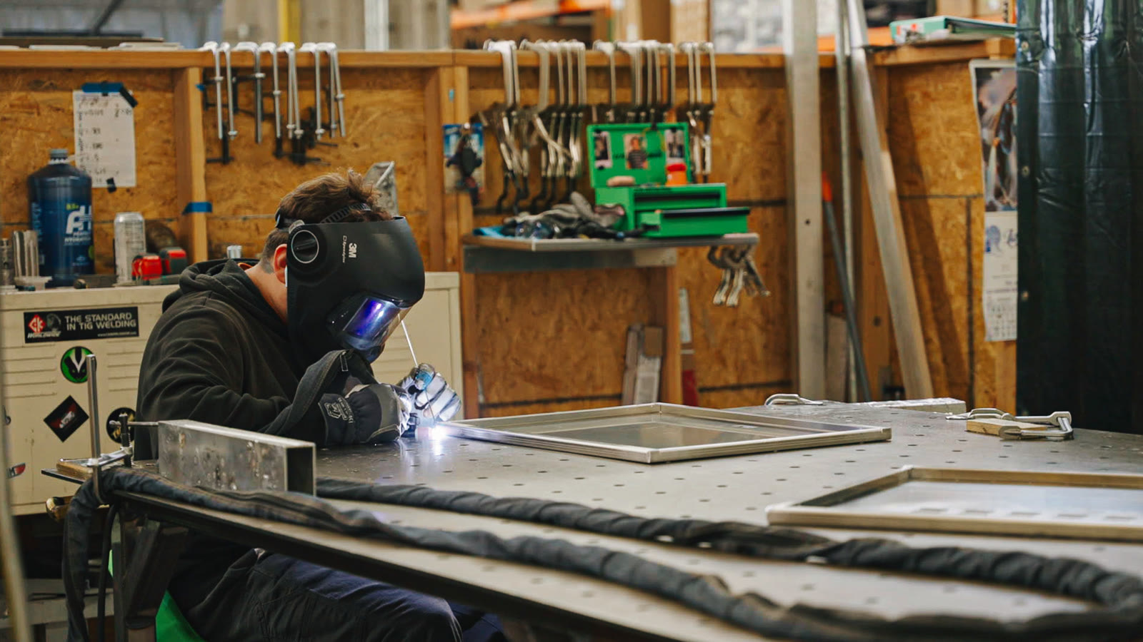 A helmeted technician works along a long silver assembly supported on the fabrication table.