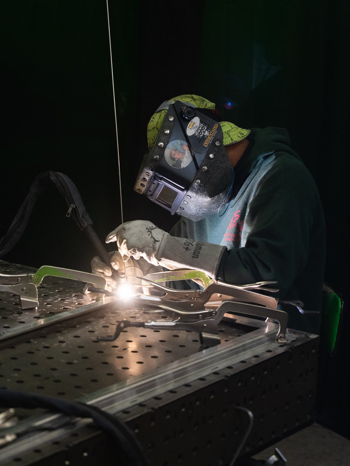 A helmeted technician works at a perforated table with sparks and a bright welding arc.