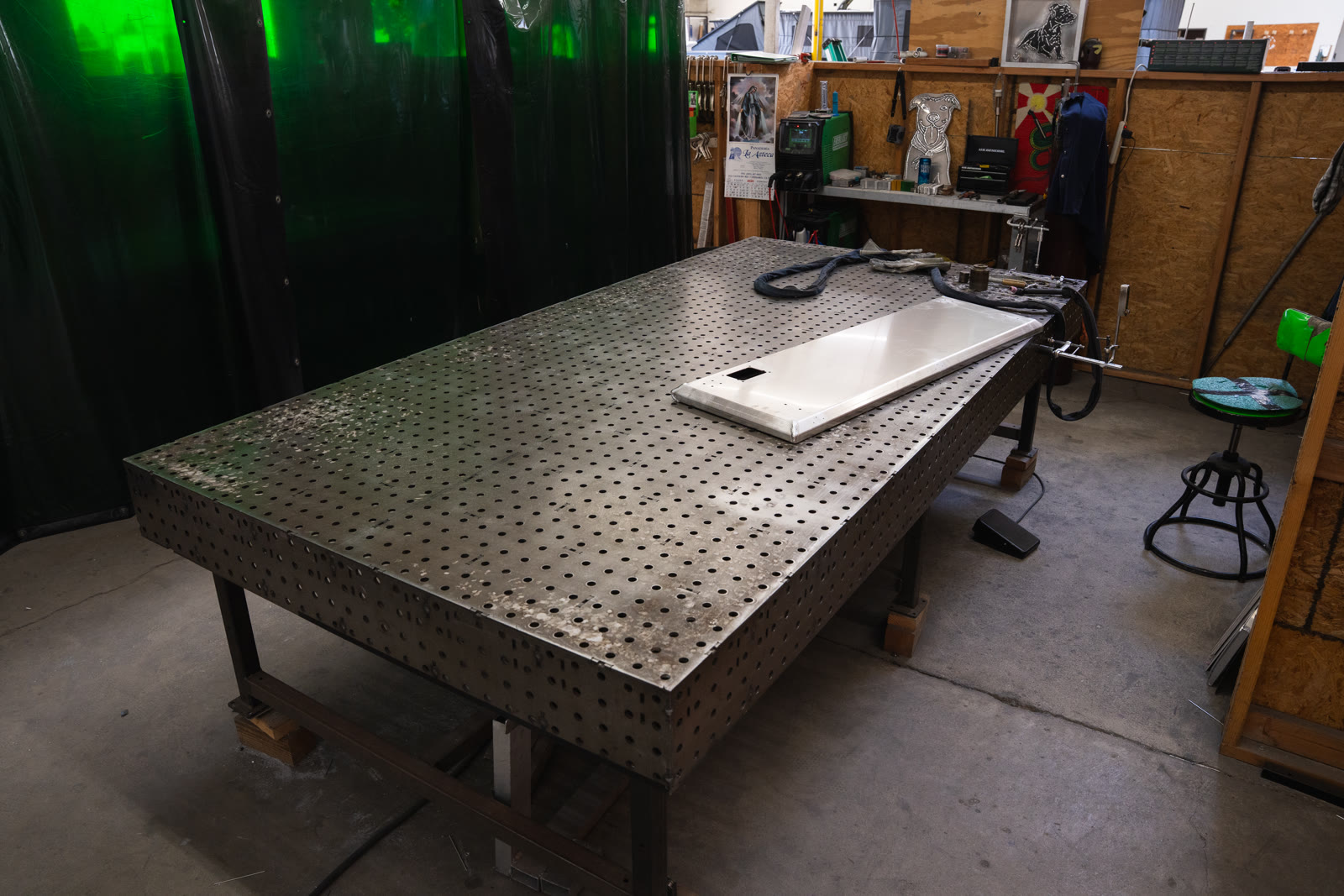 A large perforated fabrication table holds a rectangular frame and smaller silver pieces beneath green welding curtains.