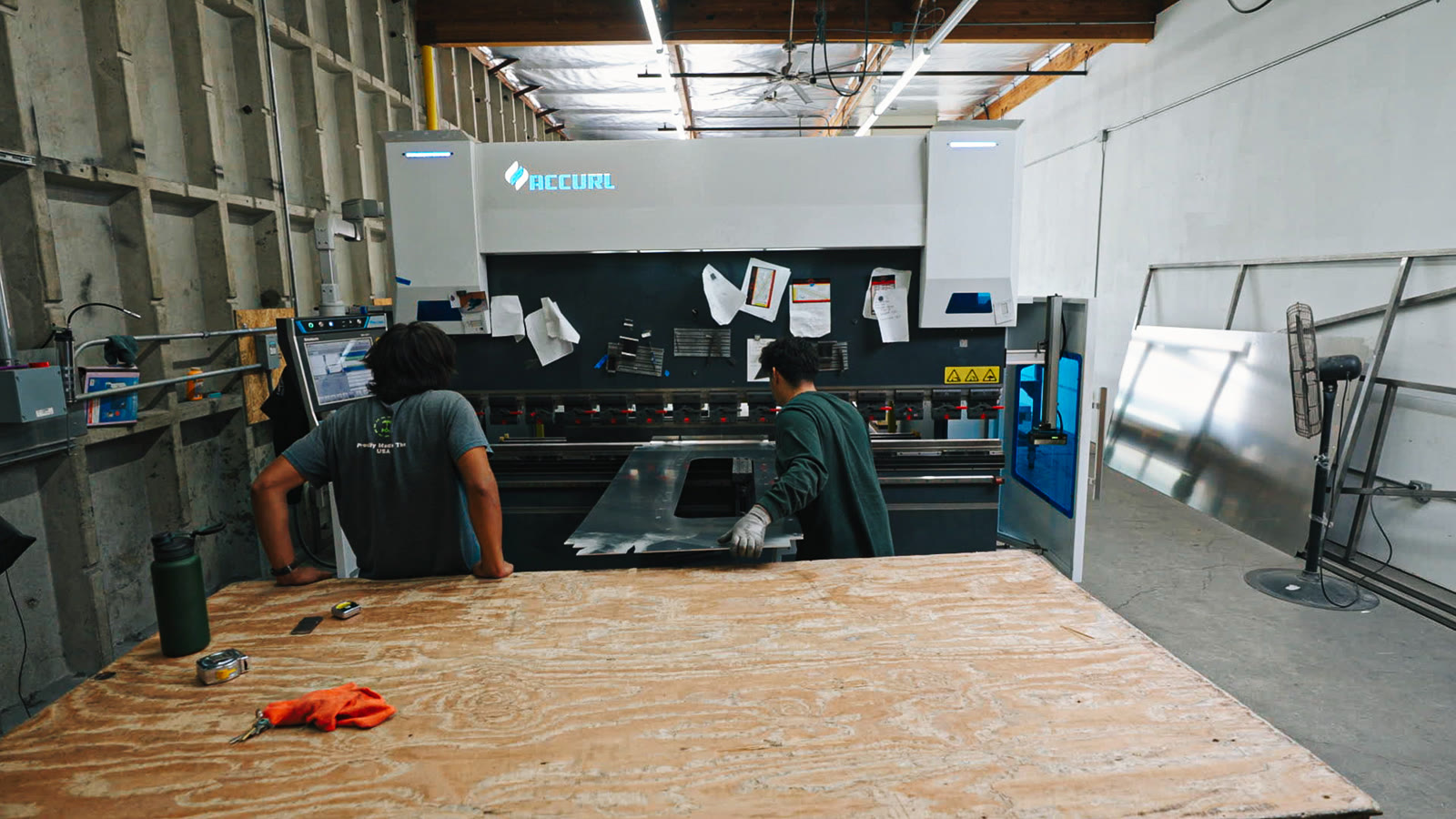 A plywood-topped table fills the foreground with workers and the bending machine behind it.