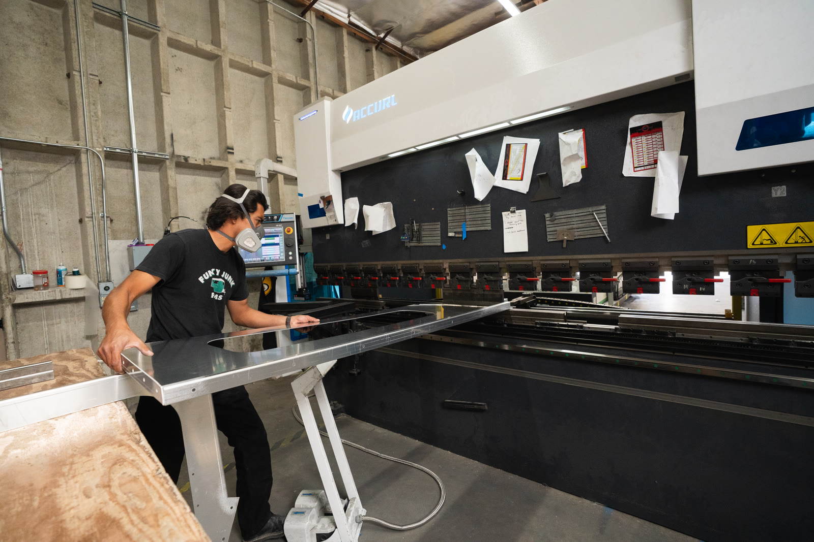 A worker carries a wide angular silver panel beside the press brake and plywood bench.
