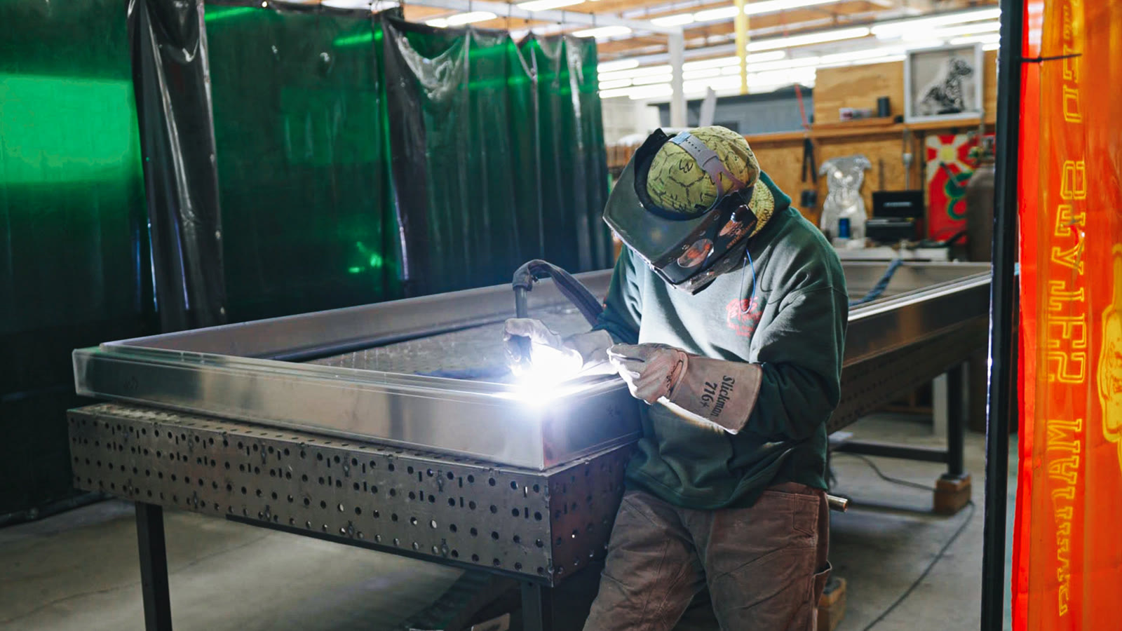 A helmeted technician leaning over a silver frame with a bright welding arc at its corner.