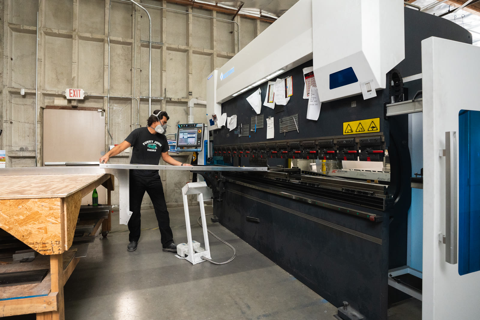 A worker carries a long silver panel between a plywood workbench and the press brake.