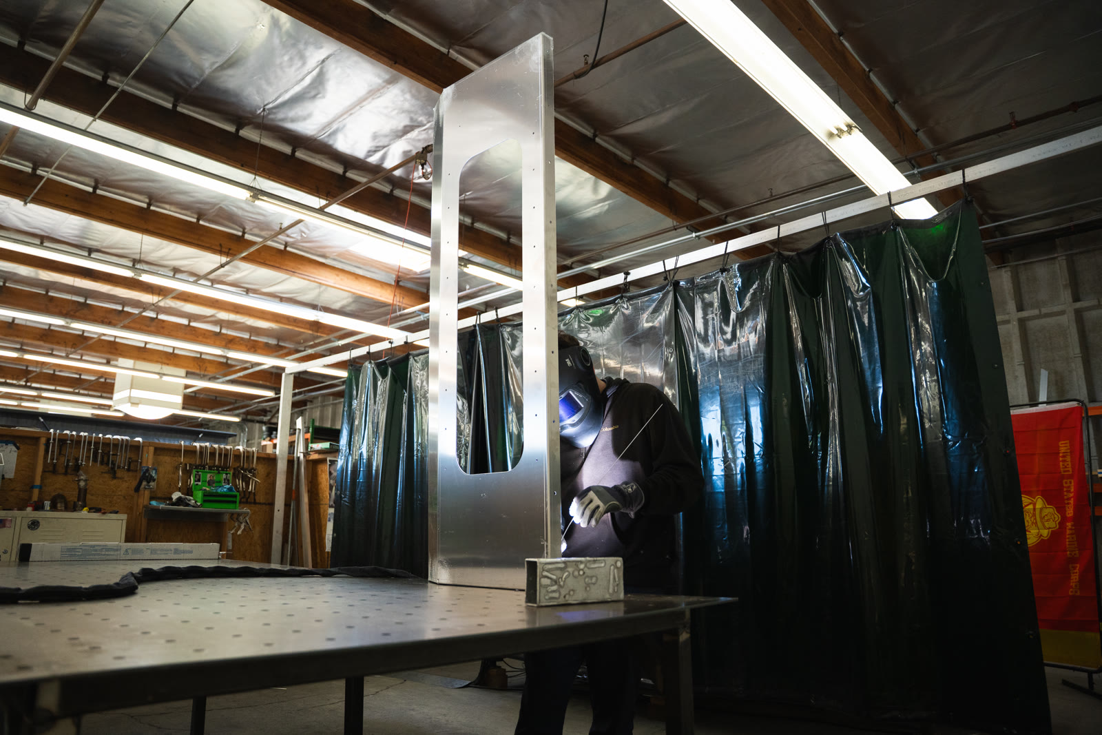Wide factory view with a tall silver frame in the foreground and a technician welding behind it.