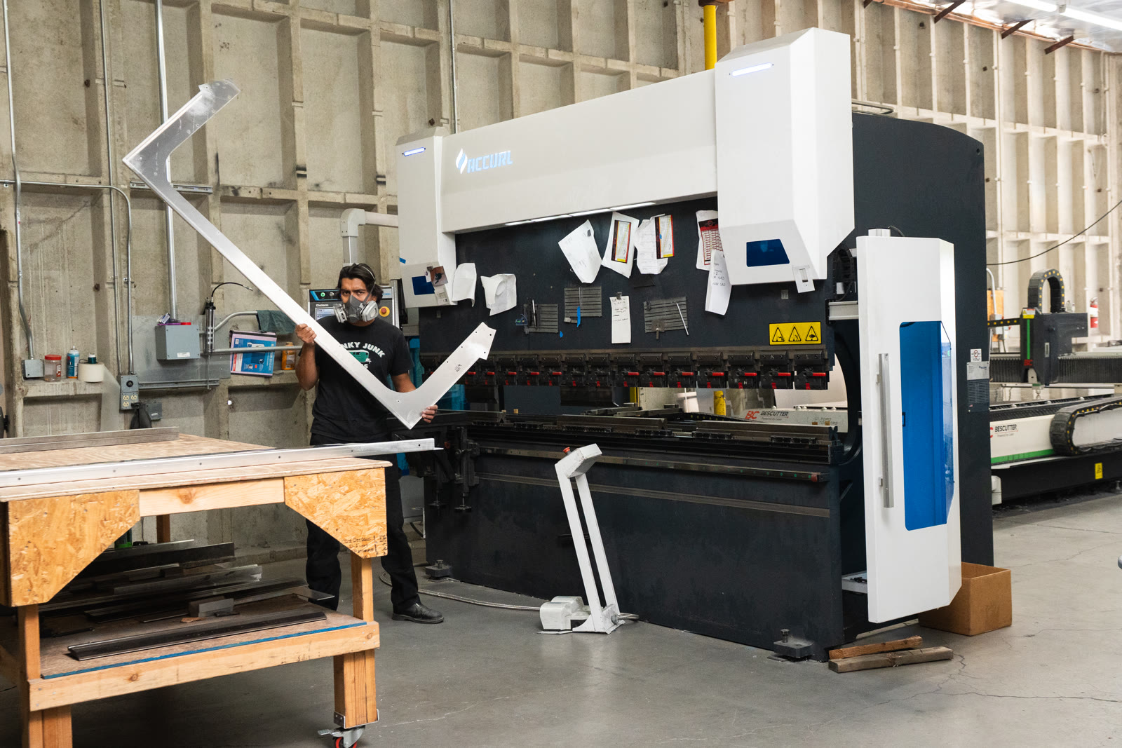 A worker holds a large angular silver frame in front of the white-and-black bending machine.