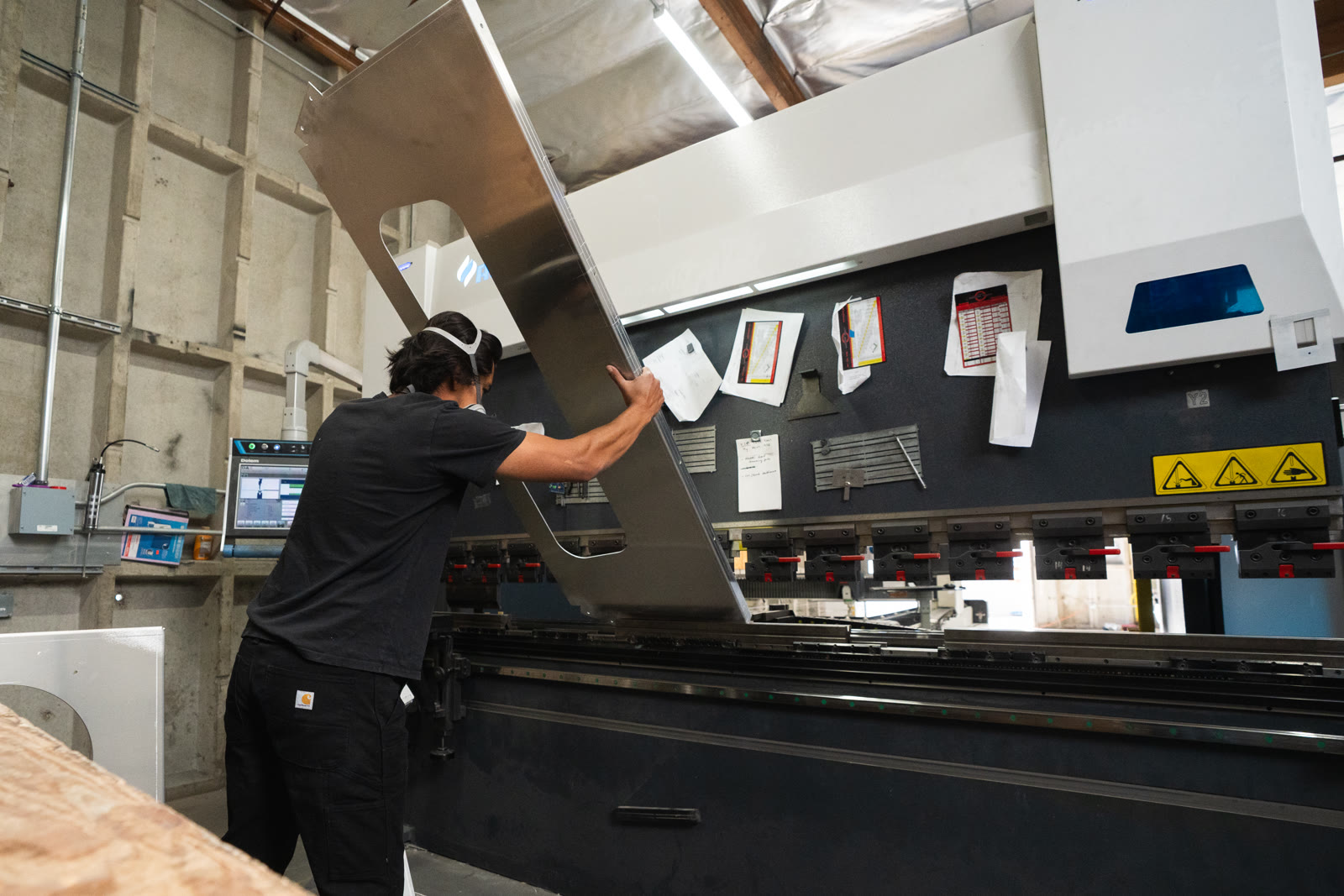 A worker lifts a broad silver panel toward the tooling of the bending machine.