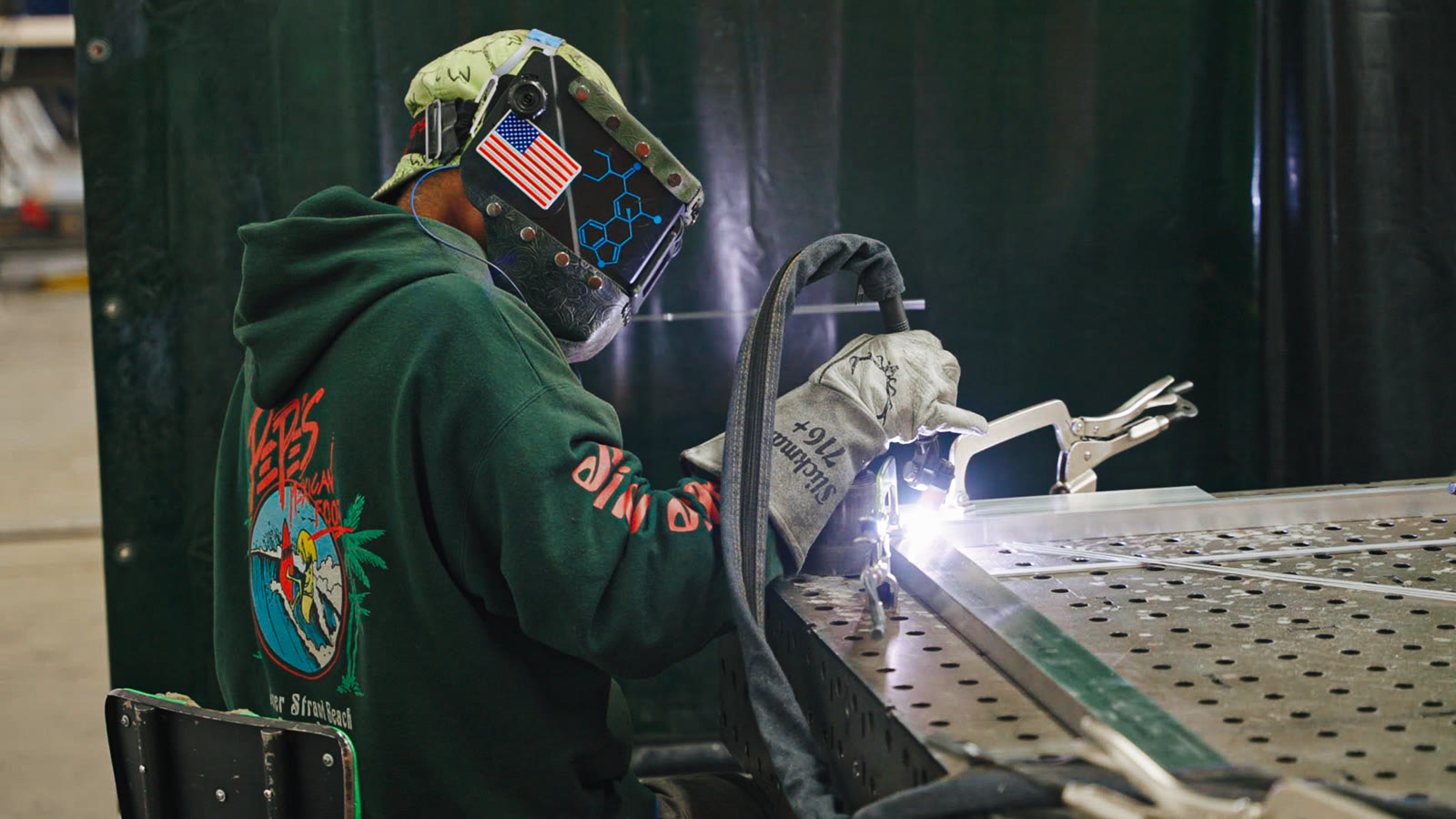 A helmeted technician seen from behind as a bright arc lights the silver frame on the table.