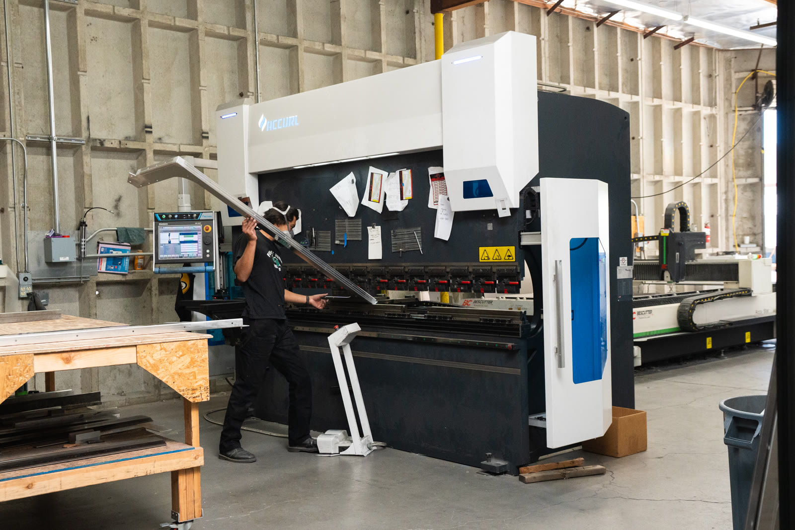 A worker raises one end of a long narrow silver piece beside the bending machine.