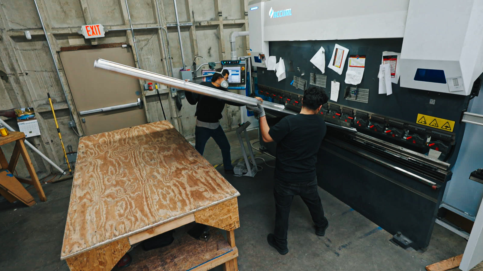 Overhead view of a worker holding a long narrow metal panel between the machine and plywood bench.