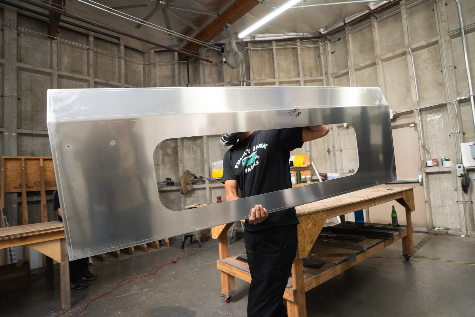 A worker carries a large silver panel with a wide rectangular opening past a wood workbench.