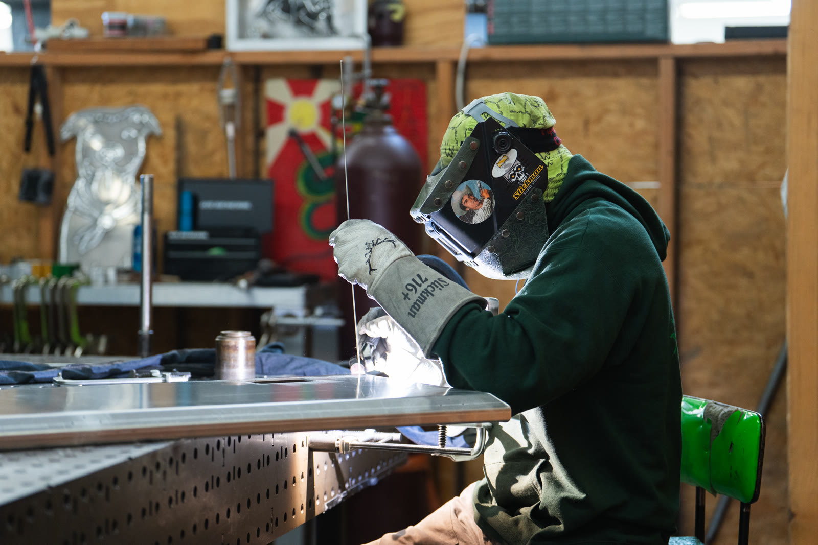 A helmeted technician holds a welding torch over a silver frame on the perforated table.